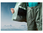 Une veste et un pantalon de ski verts avec vue sur des montagnes enneigées.