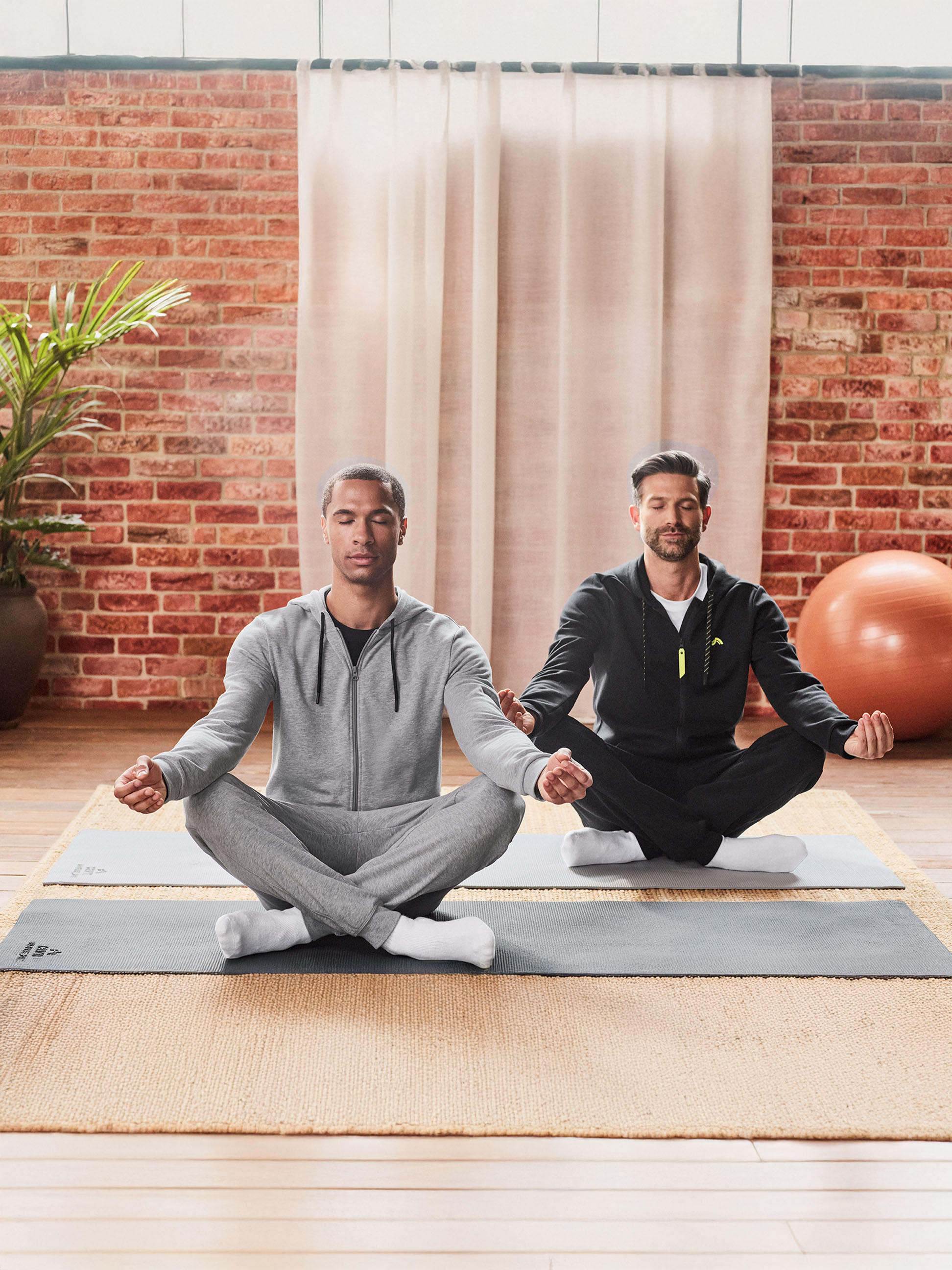 Deux hommes en survêtement méditant sur des tapis de yoga dans une pièce avec un mur de briques.