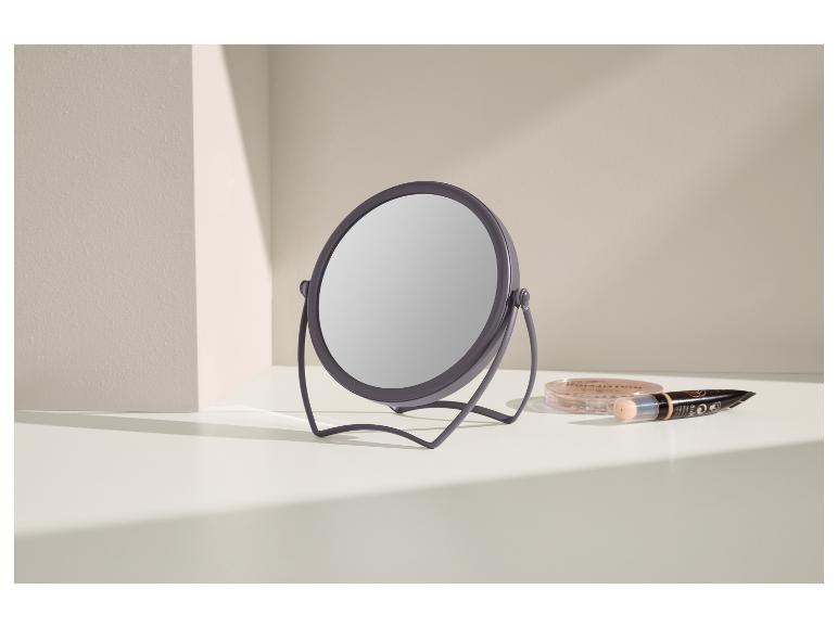 Miroir de maquillage rond avec support gris et cosmétiques sur une surface blanche.