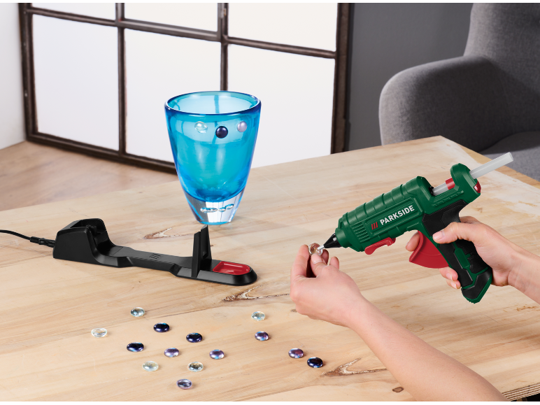 Pistolet à colle chaude décorant un vase en verre avec des gemmes sur une table en bois.