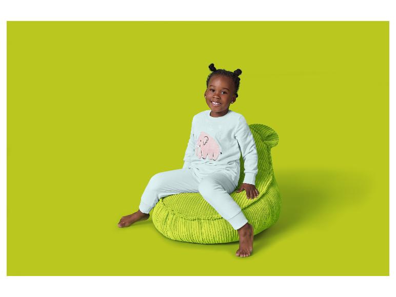 Une fille souriante assise sur un pouf vert, portant un pyjama clair avec un éléphant rose.