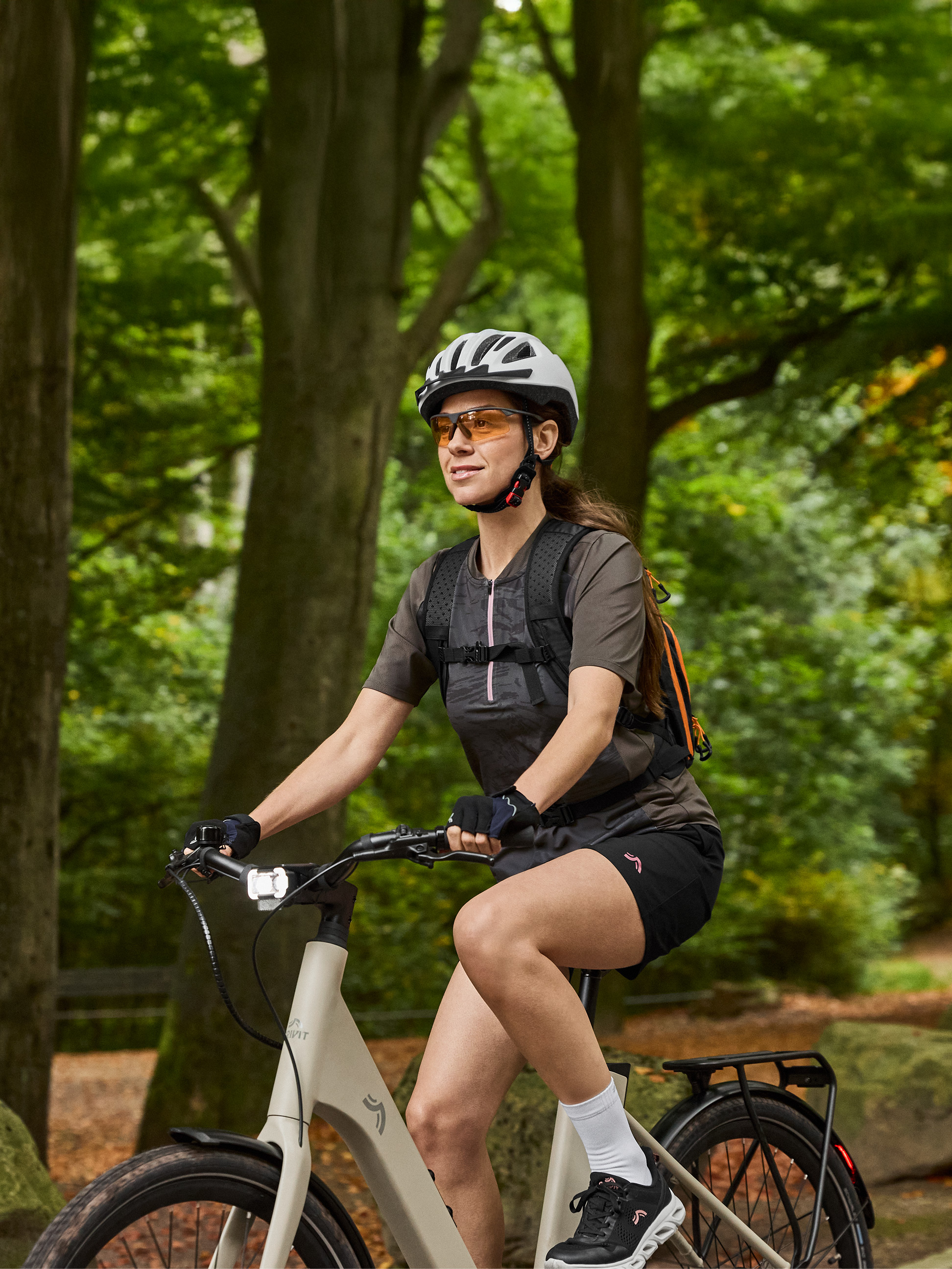 Femme à vélo électrique avec casque, lunettes de soleil et sac à dos dans une forêt.
