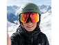 Personne avec casque et lunettes de ski, reflétant les montagnes enneigées, souriant par une journée ensoleillée.