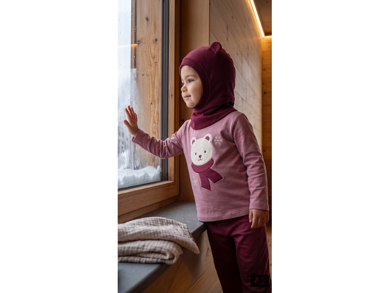 Enfant en cagoule bordeaux et haut rose avec un ours polaire, regardant par la fenêtre.