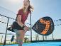Femme jouant au padel avec une raquette noire et orange sur un court bleu.