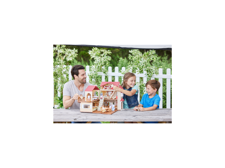 Père et deux enfants jouant avec une grande maison de poupées sur une table en bois à l'extérieur.