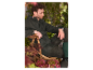 Un homme assis sur une chaise en bois dans un jardin, portant une veste vert foncé et des bottes.