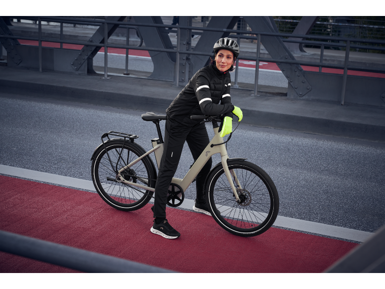 Femme en tenue de cyclisme noire et gants jaunes avec un vélo électrique sur un pont.