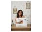 Femme souriante à une table avec une machine à coudre blanche, mur à motif géométrique en arrière-plan.