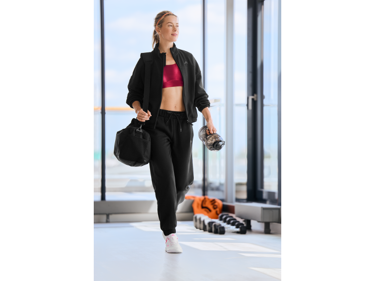 Femme en survêtement noir et brassière de sport rouge, portant un sac de sport et une gourde.