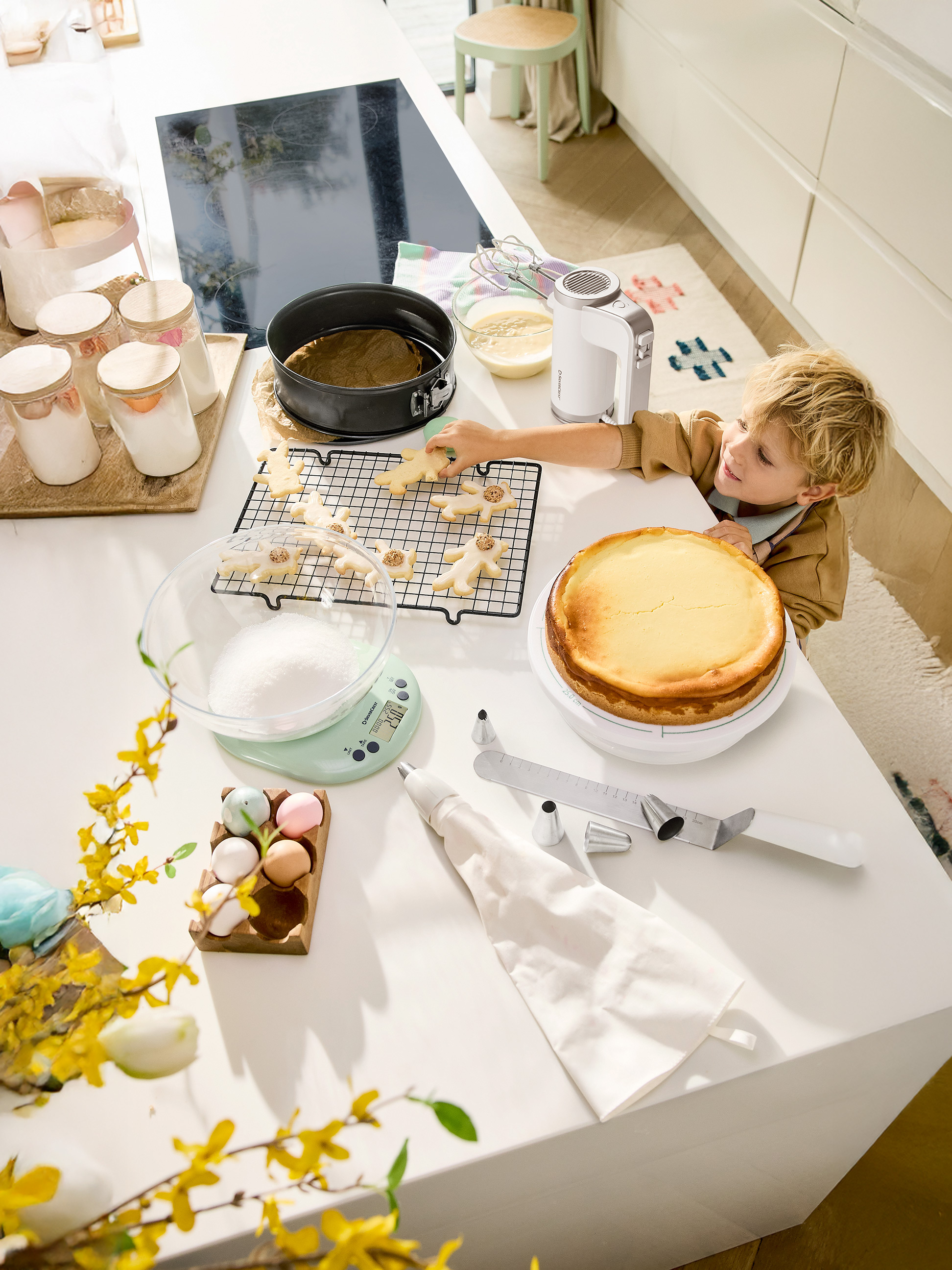 Un enfant décore des biscuits de Pâques et un cheesecake sur un comptoir de cuisine avec des ustensiles de pâtisserie.