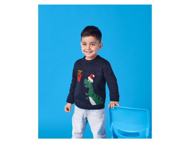 Un garçon souriant porte un pull de Noël dinosaure et un jean, appuyé sur une chaise.