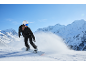 Une snowboardeuse en veste et pantalon de ski noirs, casque blanc et lunettes de ski bleues dévale une montagne enneigée.