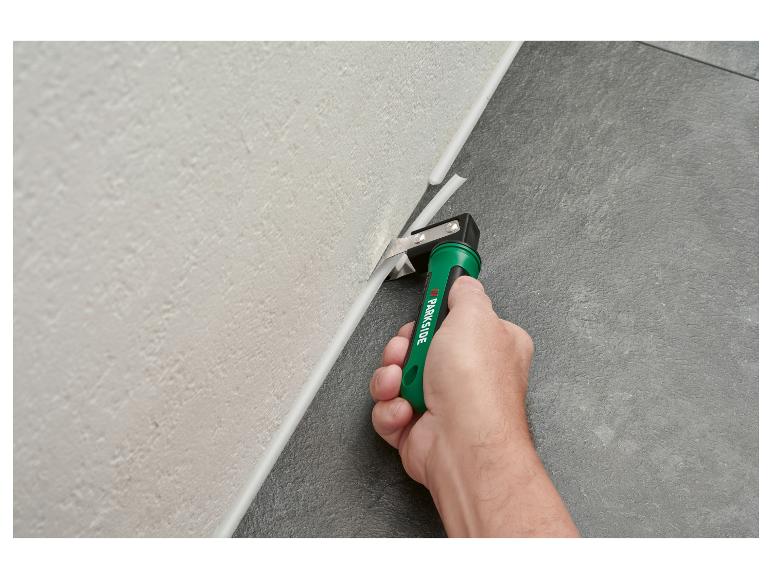 Main coupant un joint de mastic blanc entre un mur clair et un sol foncé avec un outil vert.