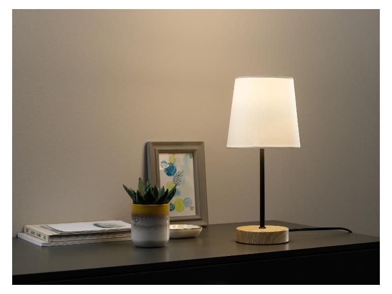 Une lampe de table avec un abat-jour blanc, une plante dans un pot jaune et des livres sur un dessus de table foncé.