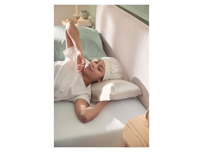 Femme dort dans un lit avec un bonnet de nuit et une taie d'oreiller en soie, le soleil éclaire son visage.