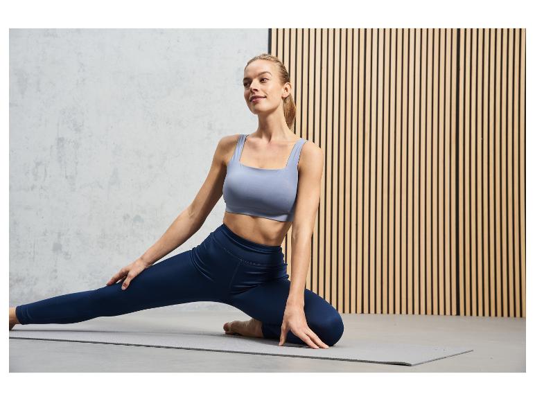 Une femme portant un soutien-gorge de sport bleu et un legging fait du yoga sur un tapis gris.