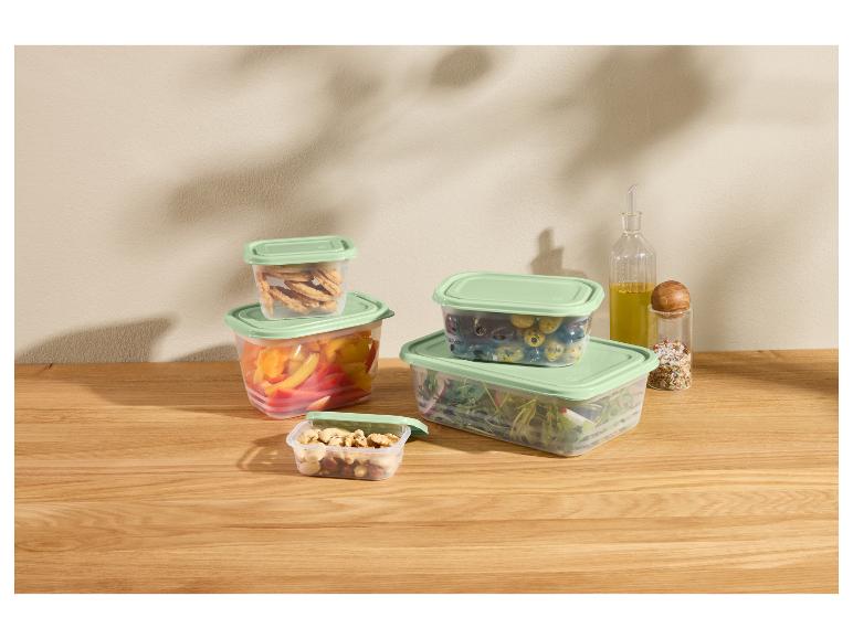 Récipients en plastique avec couvercles verts remplis de légumes, noix et biscuits sur une table en bois.