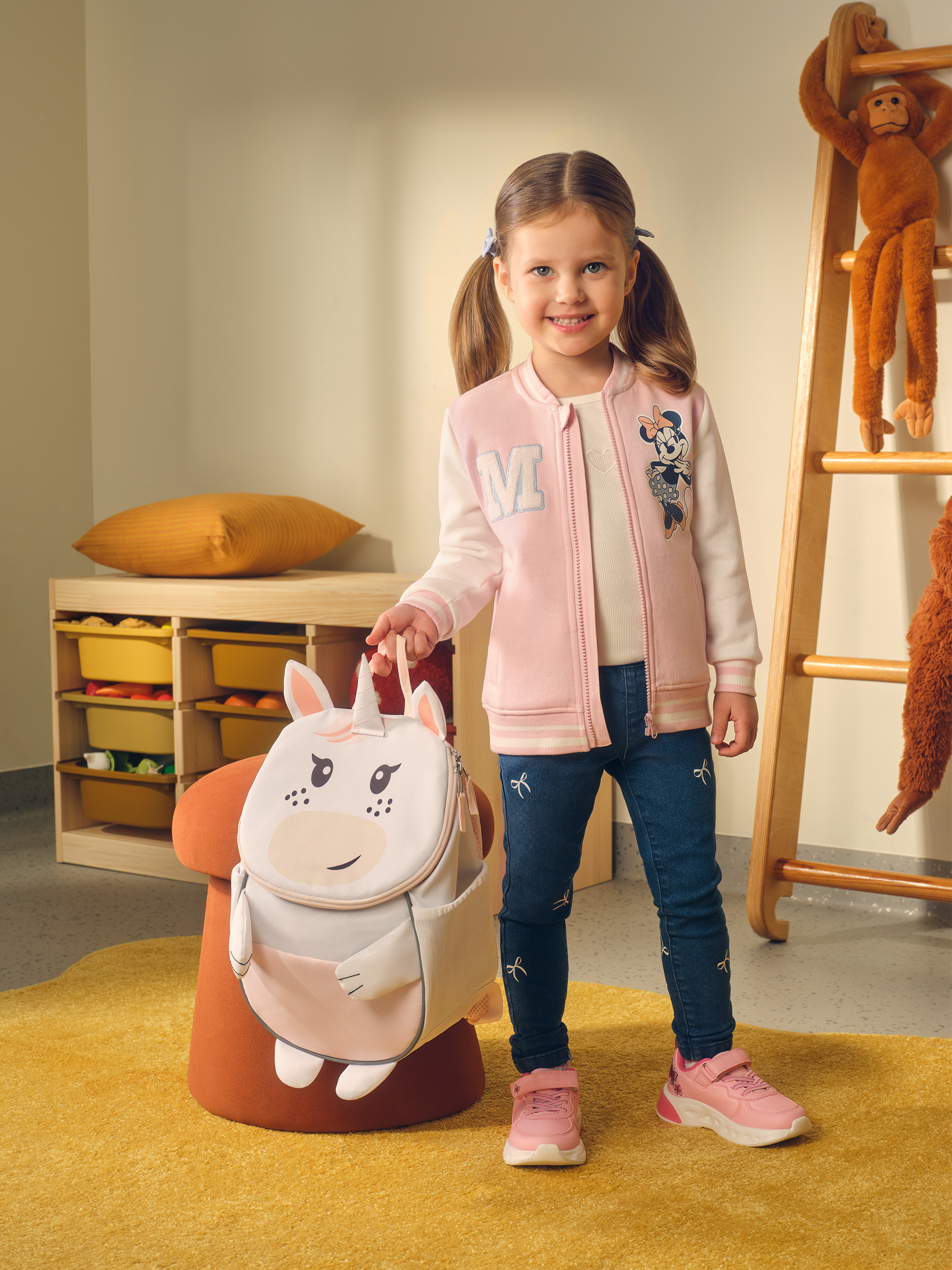 Petite fille souriante avec un sac à dos licorne, vêtue d'une veste rose et d'un jean.