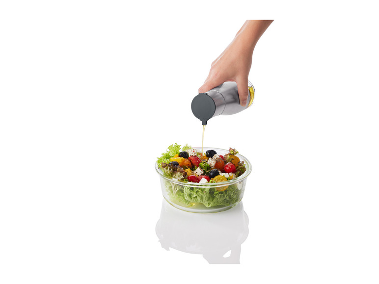 Une main verse de l'huile d'une bouteille sur une salade dans un bol en verre.