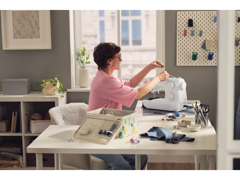 Femme cousant sur une machine à coudre, entourée d'accessoires de couture et de boîtes de rangement.