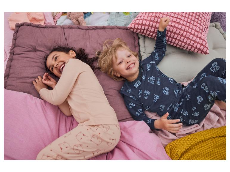 Des enfants rient, allongés dans un lit avec des draps aux couleurs pastel et des motifs de chats et de tracteurs.