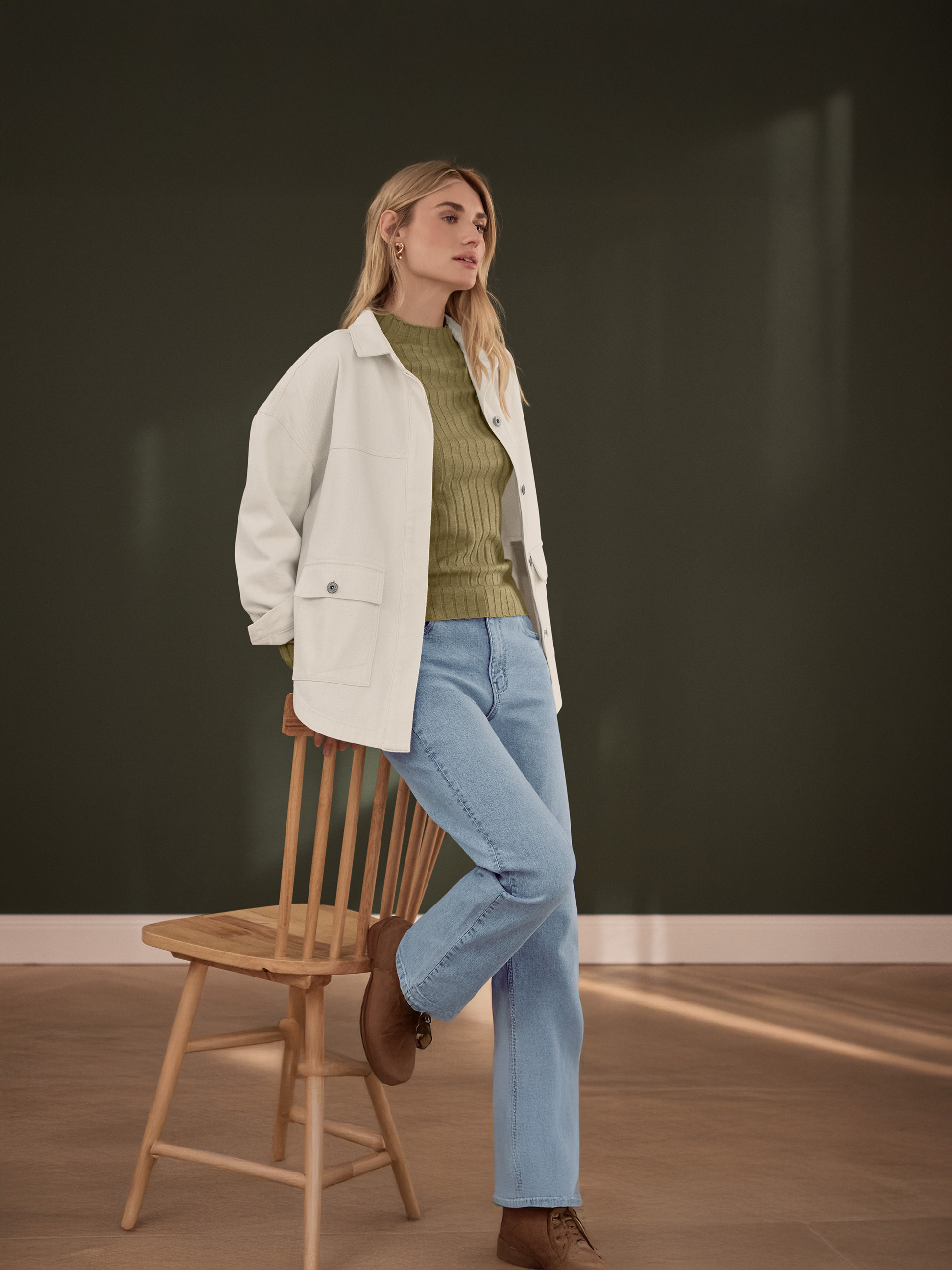 Femme en veste blanche, haut côtelé vert et jean bleu clair, appuyée sur une chaise en bois.