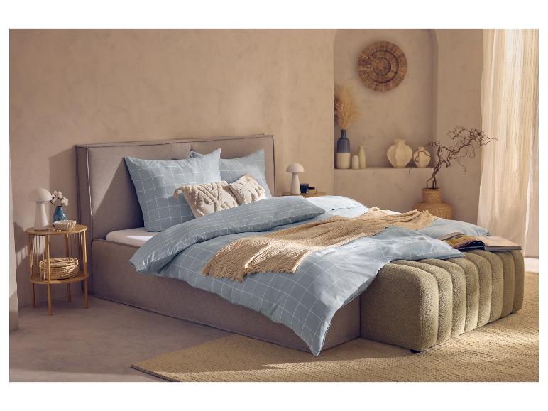 Chambre avec parure de lit à carreaux bleu clair, cadre de lit et pouf marron clair, tapis clair.