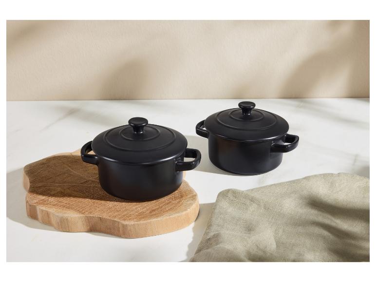 Deux mini cocottes en céramique noire avec couvercles, l'une sur une planche en bois, sur une surface blanche.