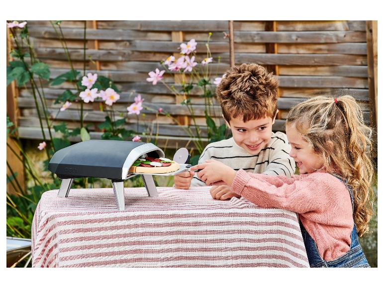 Deux enfants préparent une pizza jouet dans un four extérieur sur une nappe rayée.