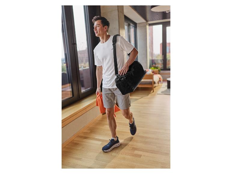 Homme en t-shirt blanc et short, avec un sac de sport et une serviette, marchant à l'intérieur.