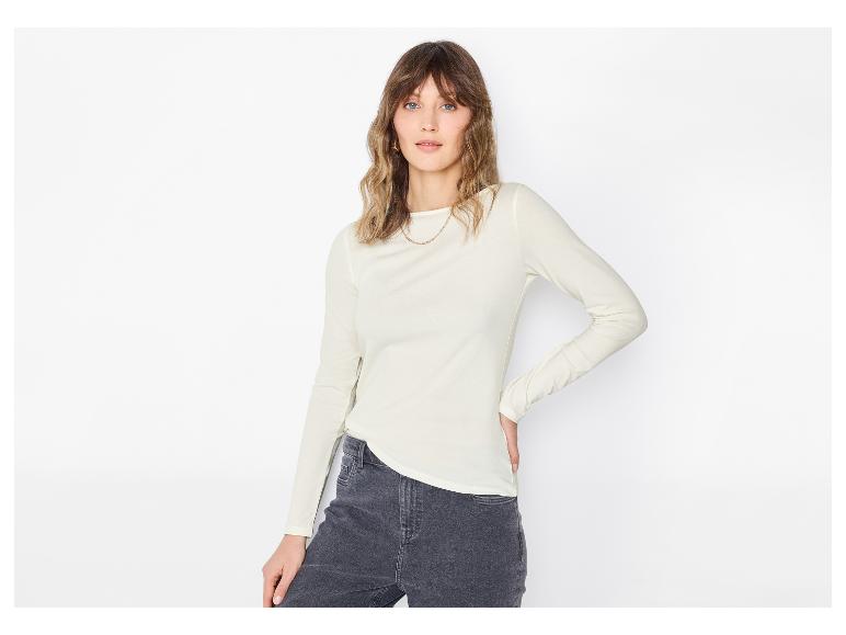 Femme en blouse claire à manches longues et jean foncé, posant sur fond blanc.