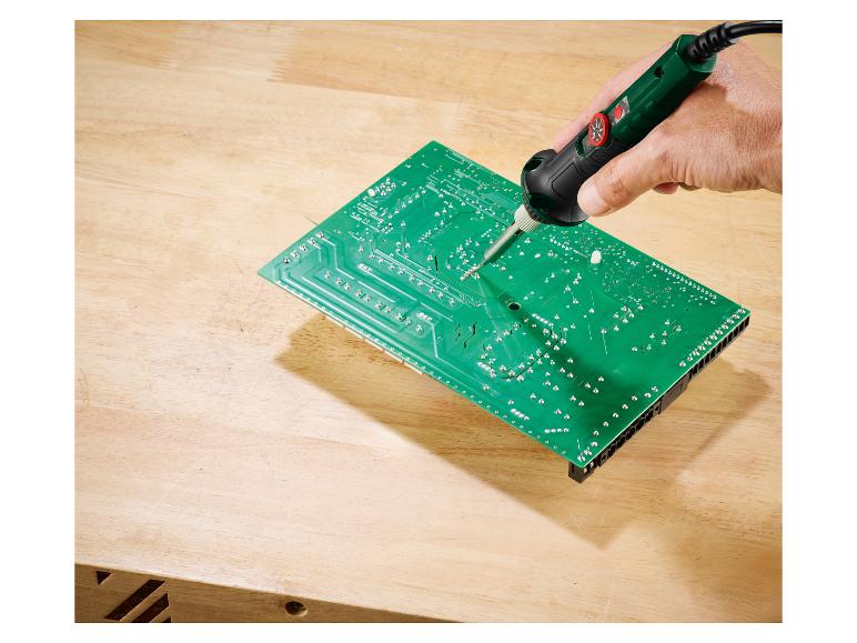 Main soudant une carte de circuit imprimé verte sur un établi en bois.