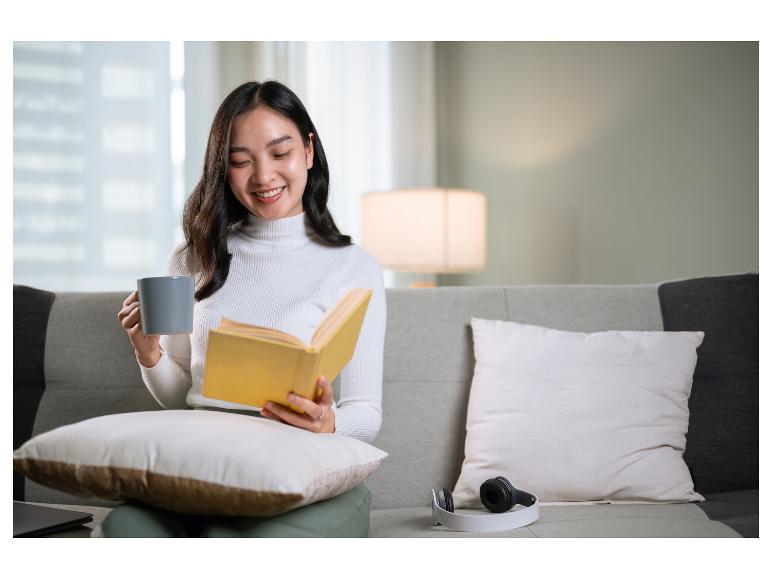 Femme souriante lisant un livre et tenant une tasse sur un canapé.