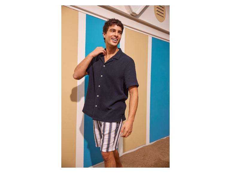 Homme en chemise bleu marine et short rayé dans un décor de plage.