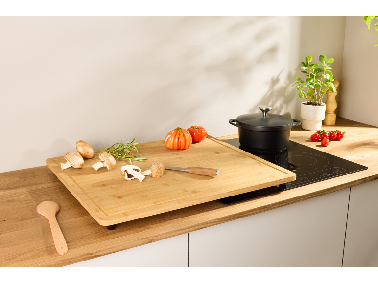 Comptoir de cuisine avec grande planche à découper en bambou, plaque à induction, casserole et légumes frais.