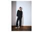Femme en sweat-shirt et pantalon de survêtement noirs, debout contre un mur blanc avec une ombre.