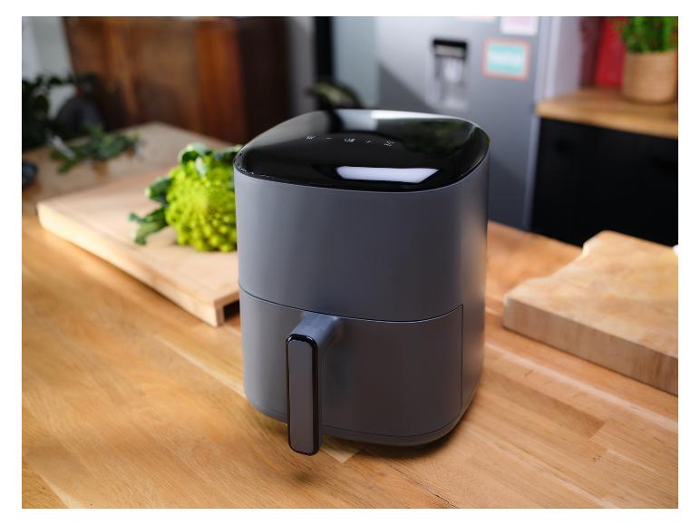 Une friteuse à air grise sur un comptoir de cuisine en bois, avec des légumes en arrière-plan.