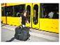 Femme portant un grand sac de voyage noir avec le logo 'SachsRAD' à côté d'un tramway jaune.