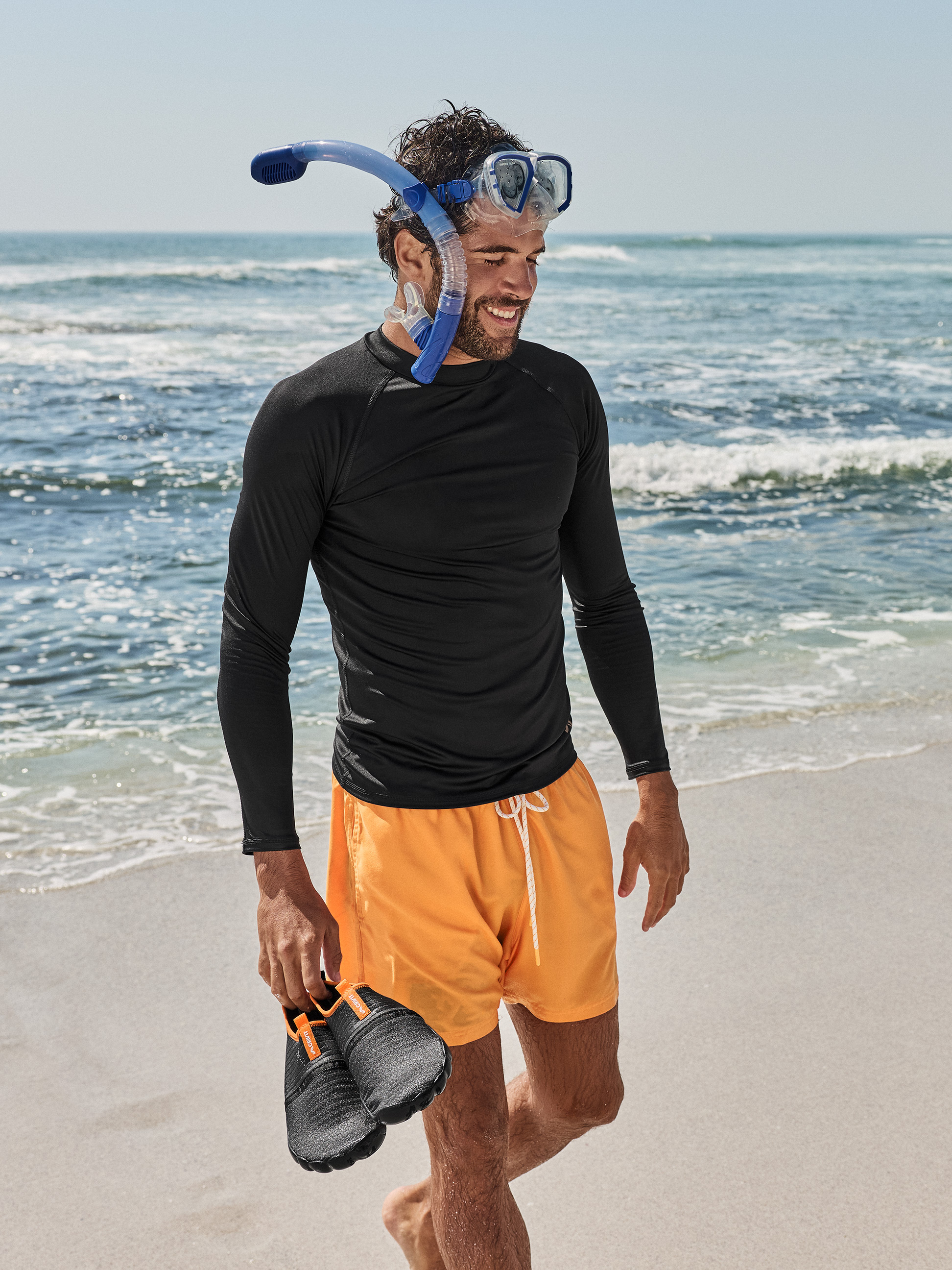 Homme en t-shirt noir à manches longues, short de bain orange, masque et tuba, tenant des chaussures d'eau sur la plage.