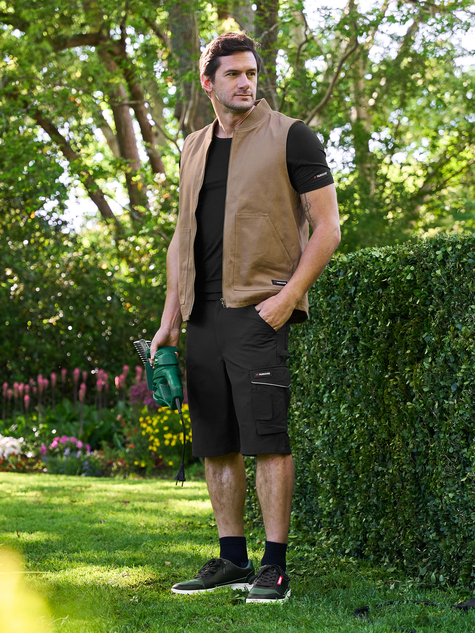 Homme en gilet et short Parkside, tenant un taille-haie électrique dans le jardin.