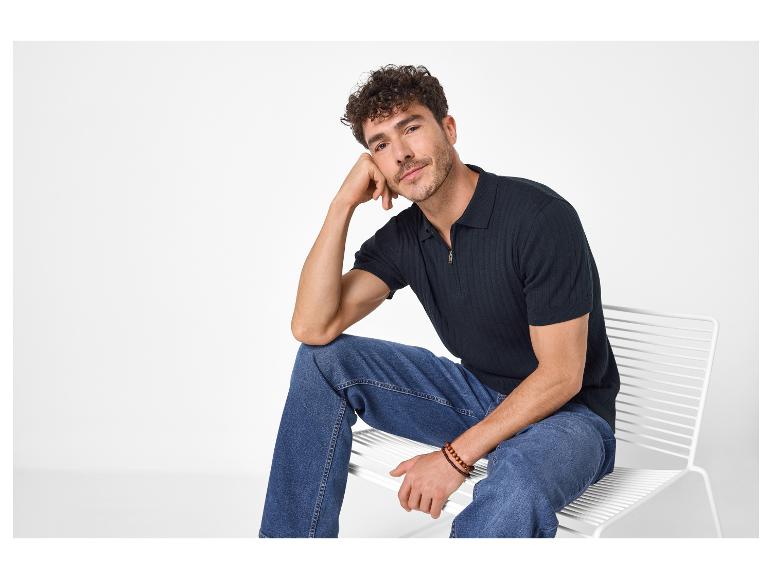 Homme en polo en maille bleu marine et jean bleu, assis sur une chaise blanche.