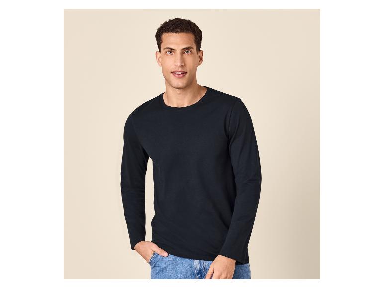 Homme portant un t-shirt noir à manches longues et un jean bleu, debout sur un fond clair.