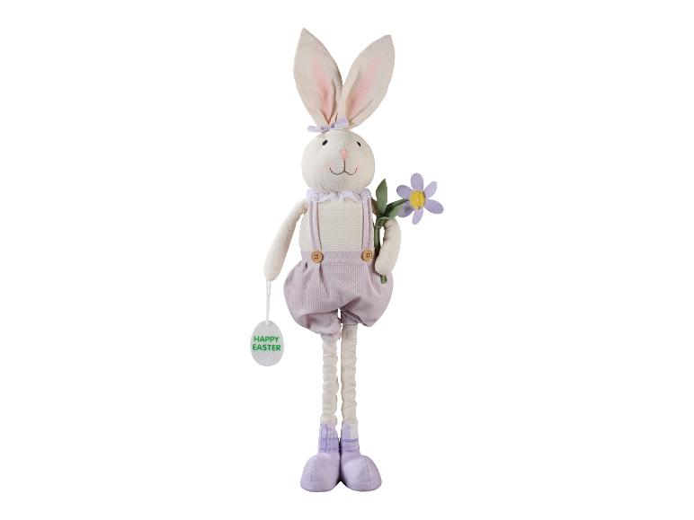 Lapin de Pâques en peluche en salopette violette, tenant une fleur et une étiquette 'Happy Easter'.