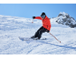 Homme en veste de ski orange et pantalon de ski noir skiant dans un paysage de montagne enneigé.
