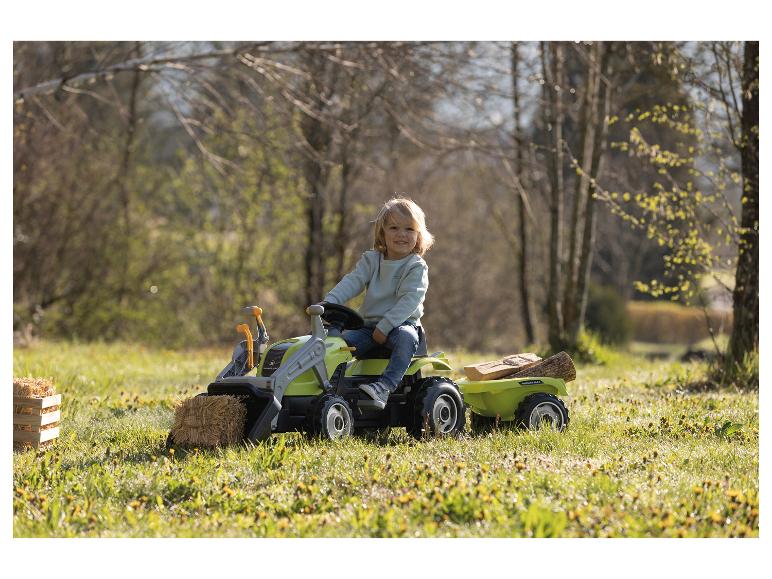 Petit enfant sur un tracteur jouet vert avec remorque et bois dans un cadre herbeux.