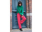 Une femme portant un pull vert, un pantalon rose et des bottes noires se tient devant une porte bleue.