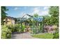 Serre avec plantes dans un jardin, à côté d'une maison et de parterres.