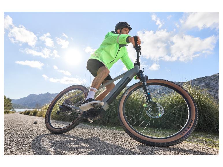 Un homme en veste vert néon conduit un vélo électrique Maxxis sur un chemin de terre.