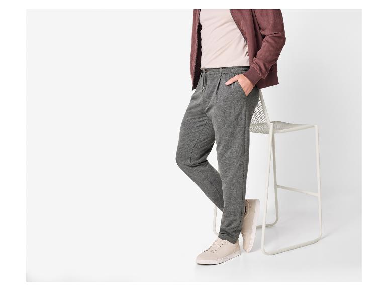 Homme en pantalon de jogging gris, t-shirt beige et veste marron, appuyé sur un tabouret.